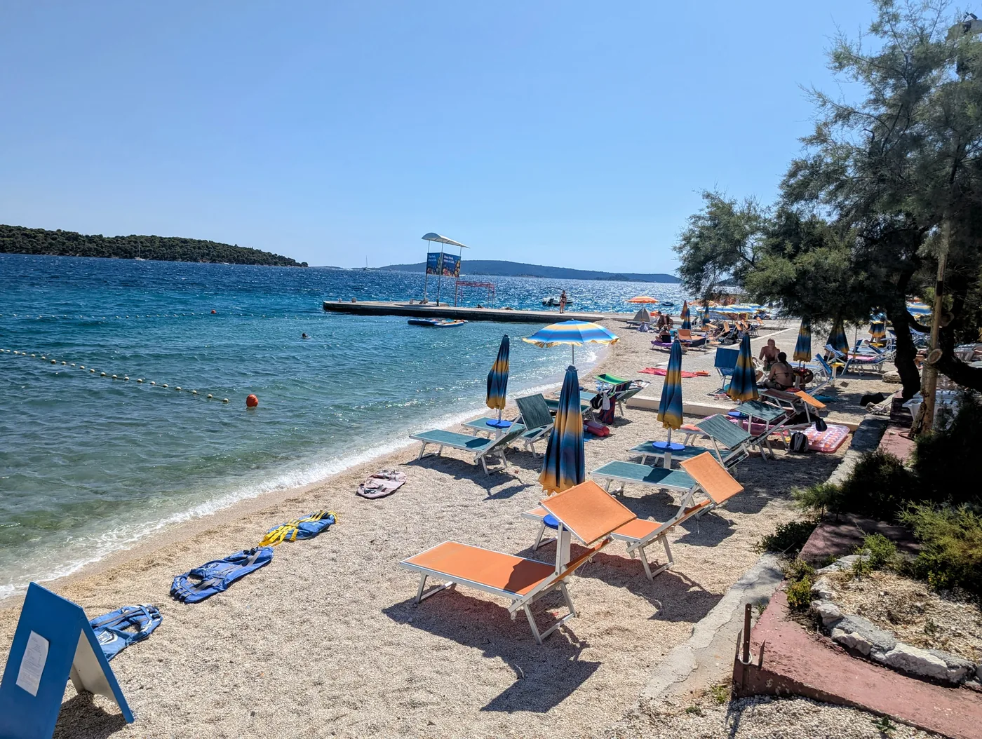 Medena beach in Seget: sunbeds and folded umbrellas along the pebble shore, clear shallow water and the islands beyond
