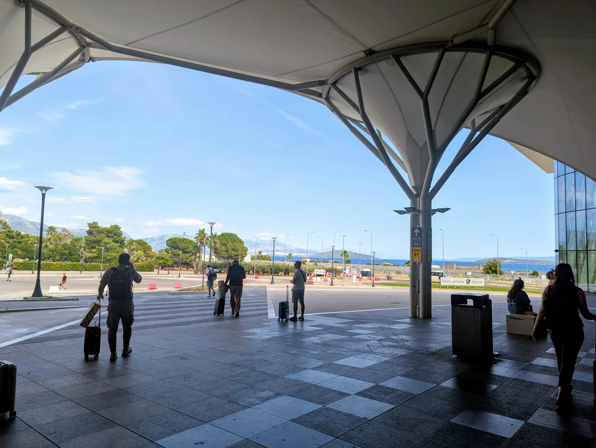 Passengers with suitcases walking out from under the curved canopy in front of Split Airport, with the sea visible beyond
