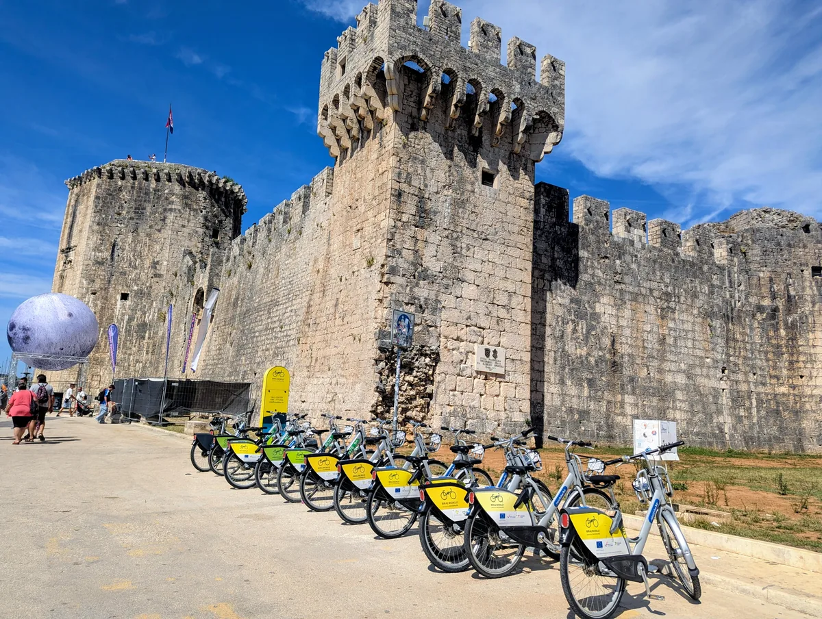 A row of hire bikes in a docking station on the promenade below the stone walls of Kamerlengo fortress in Trogir
